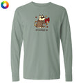 A sage green long-sleeve t-shirt with a cartoon graphic of a dog wearing sunglasses and a brown robe, holding a red book, with the text 'all booked up' below. A small multicolor color wheel icon is visible in the top-left corner.