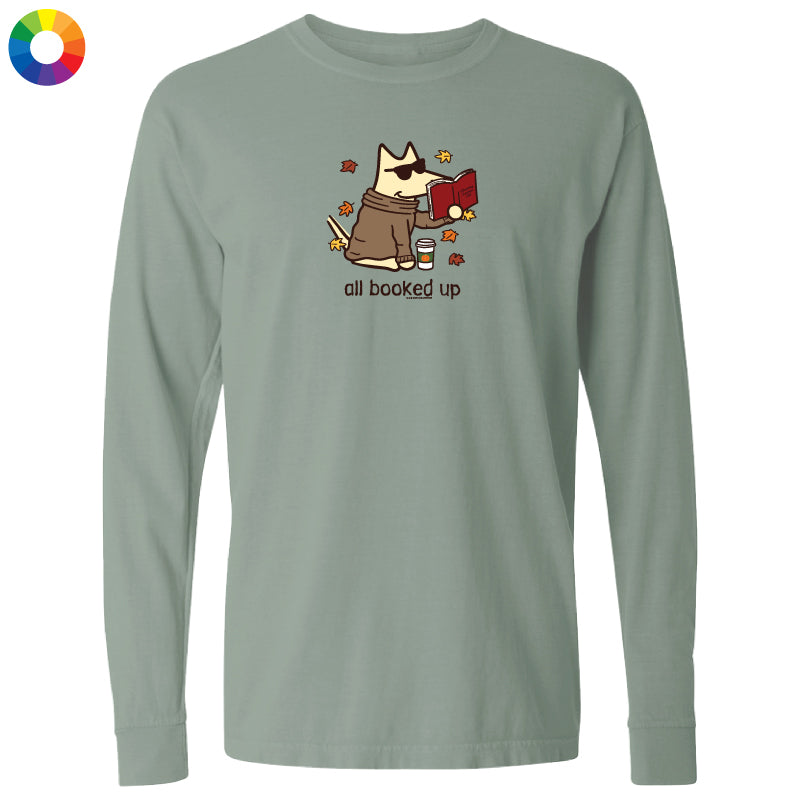 A sage green long-sleeve t-shirt with a cartoon graphic of a dog wearing sunglasses and a brown robe, holding a red book, with the text 'all booked up' below. A small multicolor color wheel icon is visible in the top-left corner.
