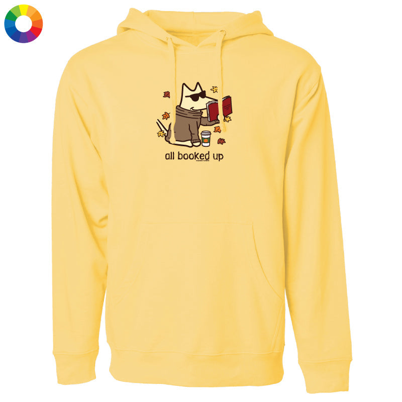 A pale yellow pullover hoodie with a front kangaroo pocket and a central graphic of a dog wearing sunglasses and a brown jacket, reading a red book, with the text 'all booked up' below. A small multicolor color wheel icon is in the top-left corner.