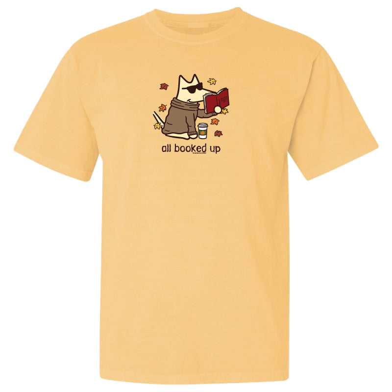 A pale yellow short-sleeve crewneck t-shirt featuring a cartoon graphic of a dog in a brown robe reading a red book, with the text 'all booked up' printed below.