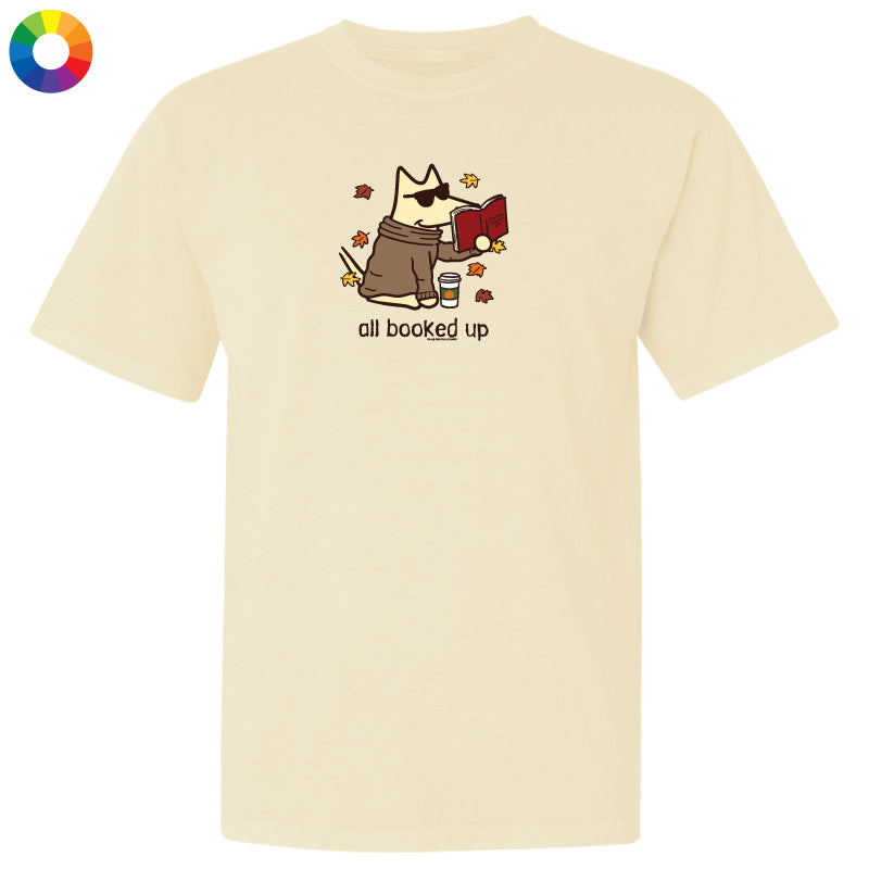 A cream-colored short-sleeve t-shirt with a cartoon graphic of a dog wearing sunglasses and reading a red book, with the text 'all booked up' below. A small multicolor color wheel icon is in the top-left corner.