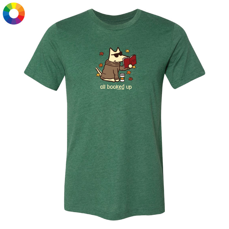 A heather green short-sleeve t-shirt with a cartoon graphic of a dog wearing sunglasses and holding a red book, with the text 'all booked up' below. A small multicolor color wheel icon is in the top-left corner, set against a plain white background.