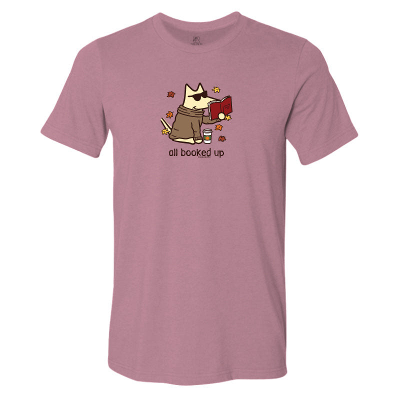 A heathered dusty rose short-sleeve t-shirt with a cartoon graphic of a dog wearing a trench coat and holding a red book, surrounded by autumn leaves, with the text 'all booked up' printed below.