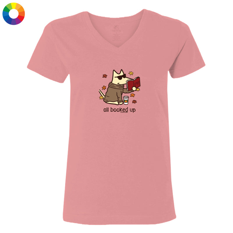 A pink V-neck t-shirt with a cartoon graphic of a dog wearing sunglasses and a trench coat while reading a red book, with the text 'all booked up' below. A small multicolor color wheel icon is in the top-left corner.