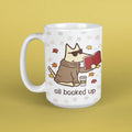 A white ceramic mug with a cartoon dog wearing sunglasses and a brown hoodie, reading a red book with autumn leaves and the text 'all booked up' on a yellow background.