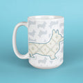 A white ceramic mug featuring a Corgi silhouette filled with a floral pattern in beige and blue, set against a background of light gray Corgi silhouettes. The mug is photographed on a solid light blue background.