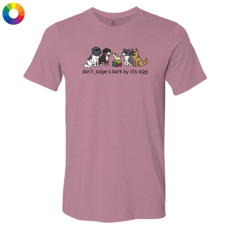 A dusty rose short-sleeve t-shirt with a graphic of four dogs of different sizes sitting together with a small rainbow-colored object, and the text 'don't judge a bark by its size' printed below. A small multicolor color wheel icon is in the top-left corner.