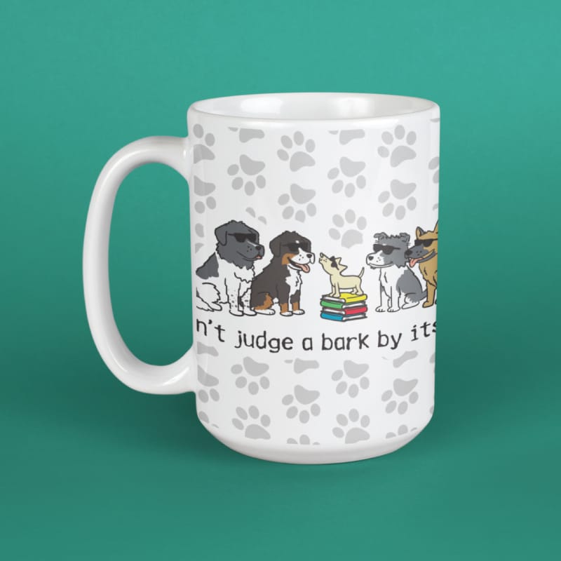 A white ceramic mug with a handle, featuring a printed design of various dog breeds sitting on books with the text 'Don't judge a bark by its.