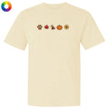 A cream-colored short-sleeve t-shirt with a horizontal row of small autumn-themed icons printed on the chest, including a paw print, maple leaf, dog, pumpkin, and sunflower. A circular multicolor color wheel icon is visible in the top-left corner.