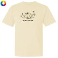 A cream-colored short-sleeve t-shirt with a graphic of a dog looking up at falling leaves, with the text 'go with the flow' underneath. A small multicolor color wheel icon is in the top-left corner.