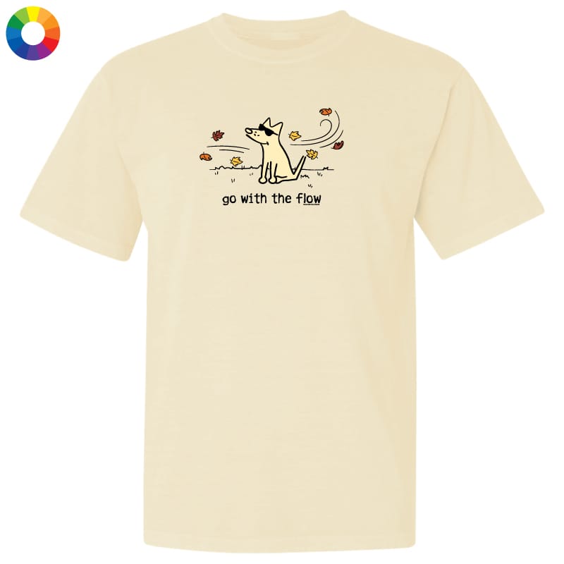 A cream-colored short-sleeve t-shirt with a graphic of a dog looking up at falling leaves, with the text 'go with the flow' underneath. A small multicolor color wheel icon is in the top-left corner.