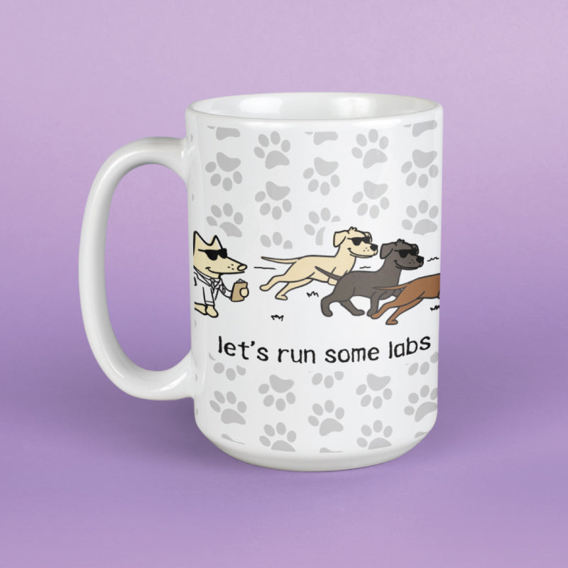 A white ceramic mug with a handle, decorated with a pattern of light gray paw prints and an illustration of three cartoon dogs wearing sunglasses, with the text 'let's run some labs' printed below. The mug is set against a solid lavender-purple background.