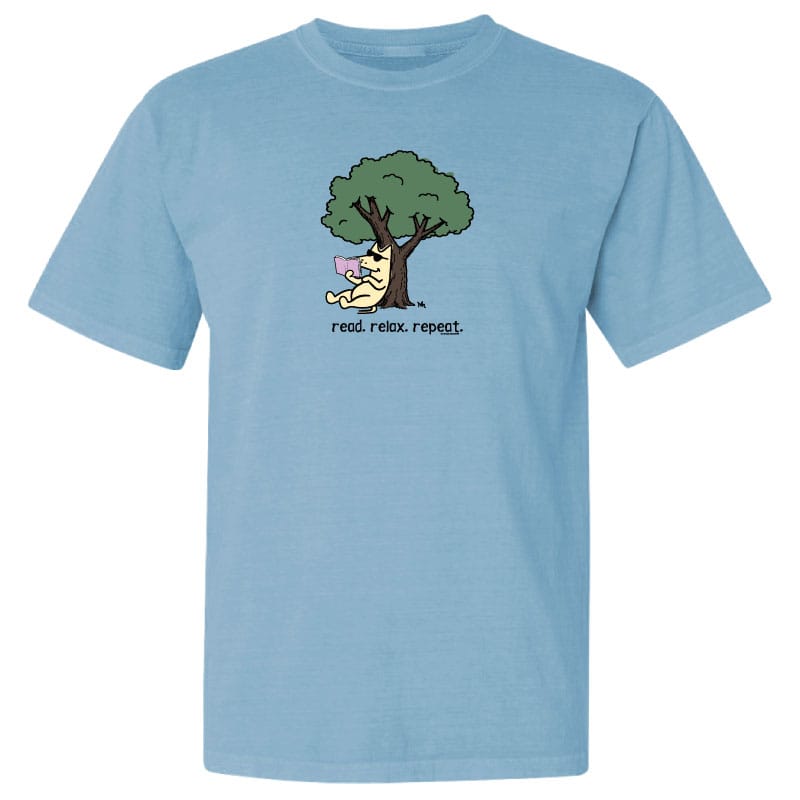 A light blue short-sleeve crewneck t-shirt featuring a cartoon graphic of a dog sitting under a tree reading a book, with the text 'read. relax.