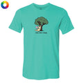 A teal short-sleeve crewneck t-shirt featuring a cartoon graphic of a yellow cat under a green tree with the text 'read, relax, repeat!' printed below. A small multicolor color wheel icon is visible in the top-left corner.