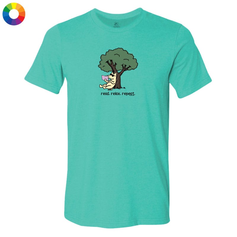 A teal short-sleeve crewneck t-shirt featuring a cartoon graphic of a yellow cat under a green tree with the text 'read, relax, repeat!' printed below. A small multicolor color wheel icon is visible in the top-left corner.