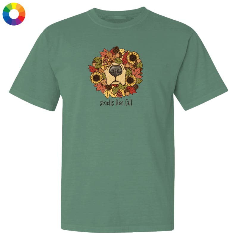 A sage green short-sleeve t-shirt with a graphic of a dog's face surrounded by autumn leaves and sunflowers, with the text 'smells like fall' underneath. A small multicolor color wheel icon is in the top-left corner.