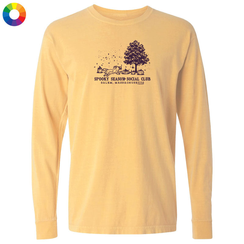 A pale yellow long-sleeve t-shirt with a dark graphic on the chest depicting a spooky season scene with a tree, haunted houses, and the text 'SPOOKY SEASON SOCIAL CLUB' and 'PALMB, MASSACHUSETTS'. A multicolor color wheel icon is in the top-left corner.