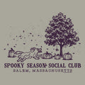 A vintage-style autumn graphic on a warm gray background, featuring a dark purple illustration of a tree, pumpkins, falling leaves, and a running dog. Text below reads 'SPOOKY SEASON SOCIAL CLUB' and 'SALEM, MASSACHUSETTS.