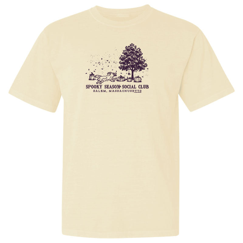 A cream-colored short-sleeve t-shirt with a dark purple graphic on the chest depicting a tree, pumpkins, and the text 'Spooky Season Social Club' and 'Salmon, Massachusetts'.