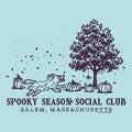 A light blue graphic print featuring a dark purple illustration of a running dog, pumpkins, a tree, and falling leaves, with the text 'SPOOKY SEASON SOCIAL CLUB' and 'SALEM, MASSACHUSETTS' below.