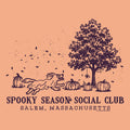 A peach-colored graphic print featuring a dark purple illustration of a running dog, pumpkins, a tree, and falling leaves, with the text 'SPOOKY SEASON SOCIAL CLUB' and 'SALEM, MASSACHUSETTS' below.