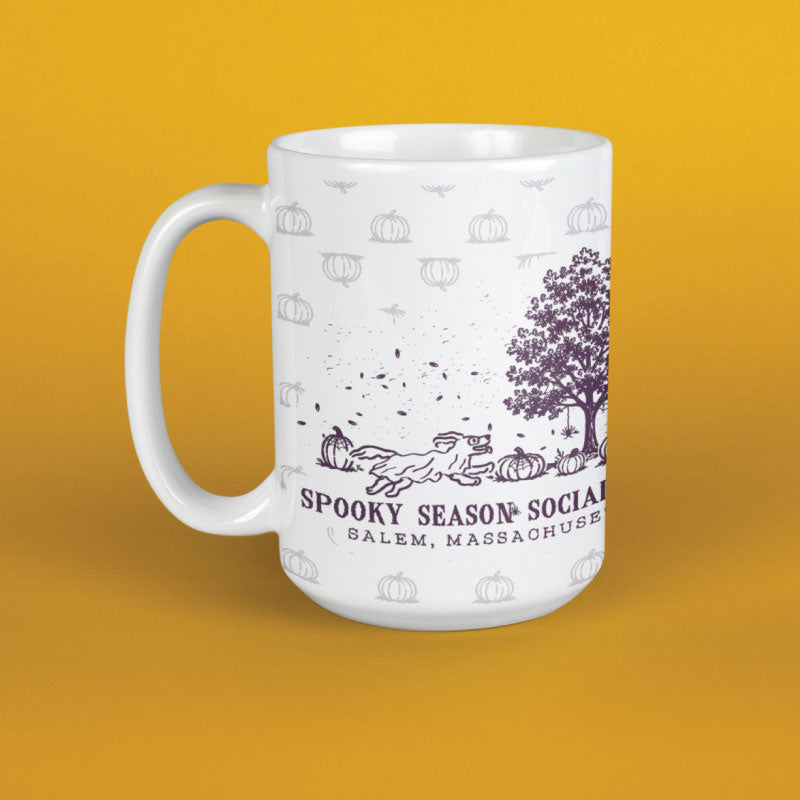 A white ceramic mug with a purple and gray autumn-themed graphic featuring pumpkins, a tree, a dog, and the text 'SPOOKY SEASON SOCIAL' and 'SALEM, MASSACHUSETTS', shown on a solid yellow background.