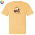 A pale yellow short-sleeve t-shirt with a graphic of a dog wearing sunglasses and a plaid scarf, next to a basket of red apples, with the text 'the dog days of fall' printed below. A small multicolor color wheel icon is in the top-left corner.
