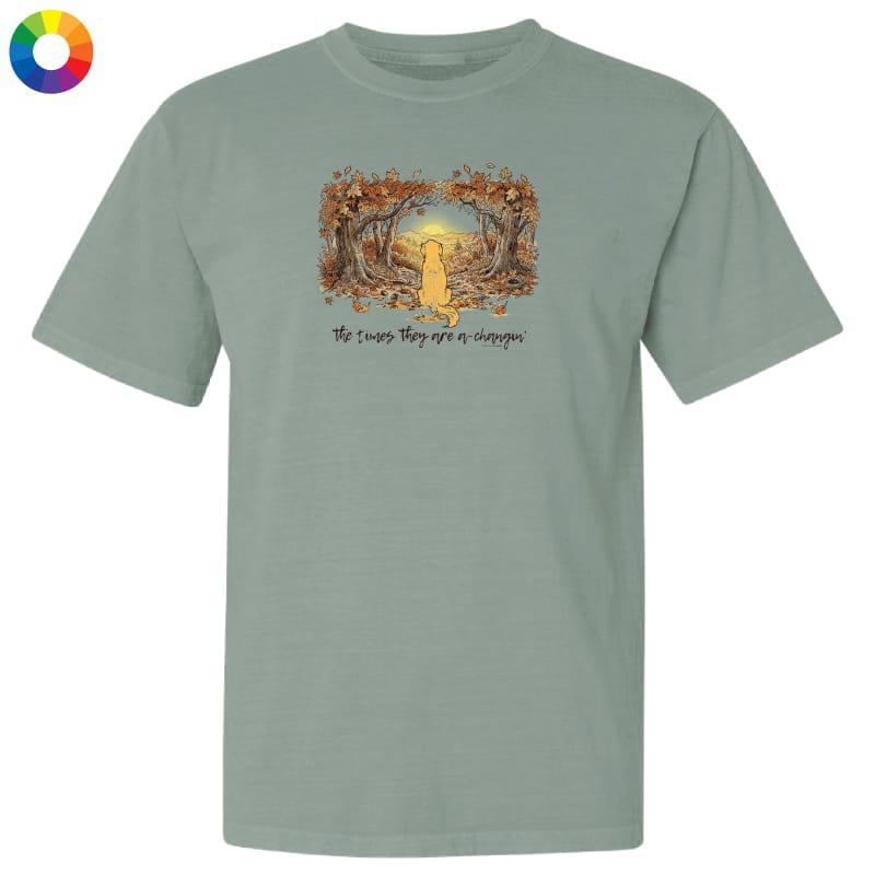 A sage green short-sleeve t-shirt with a graphic of a yellow cat sitting in a forest of trees with autumn leaves, and the text 'The cats, they see or disappear' printed below. A color wheel icon is visible in the top-left corner.