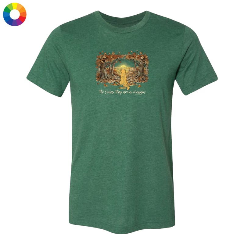 A heather green short-sleeve crewneck t-shirt with a central graphic of a yellow dog running through a forest with the text 'The cows they see a dog'. A small multicolor color wheel icon is in the top-left corner, all on a solid white background.