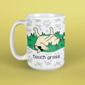 A white ceramic mug with a cartoon illustration of a cow rolling on its back in grass, with the text 'touch grass' printed below. The mug has a white paw-print pattern around the rim and is set against a solid pale yellow background.