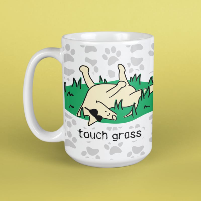 A white ceramic mug with a cartoon illustration of a cow rolling on its back in grass, with the text 'touch grass' printed below. The mug has a white paw-print pattern around the rim and is set against a solid pale yellow background.